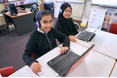 Decorative image of 2 children in a classroom with laptops and wearing headphones.