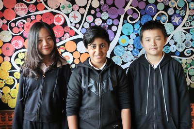 Decorative image of 3 children standing together in front of a large and colourful artwork.